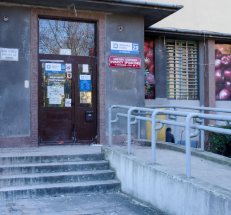 Fotografia przedstawia wejście do budynku. Przed drzwiami kilka schodów oraz pochylnia. Na drzwiach do budynku tabliczki informacyjne, na ścianie tabliczka z oznaczeniem Biblioteki oraz MOPS.