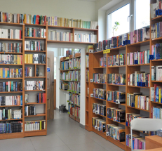 Na fotografii pomieszczenie biblioteki. Wzdłuż ścian ustawione regały z książkami w kolorze drewna. W głębi przejście do kolejnego pomieszczenia. Po prawej stronie okno, przez które wpada światło, na podłodze posadzka.