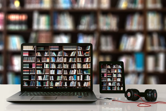 Laptop na biurku, po prawej stronie tablet i słuchawki nauszne podłączone do smartfona. Na ekranach obu urządzeń wyświetla się grafika z książkami na półkach.