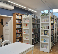 Sala biblioteczna na piętrze, szerokie wejście do pomieszczenia, wieszak na odzież w korytarzu, nad drzwiami klimatyzator. W pomieszczeniu regały z książkami, drewniane stoliki i krzesła, na półkach wystawa ręcznie robionych kartek.